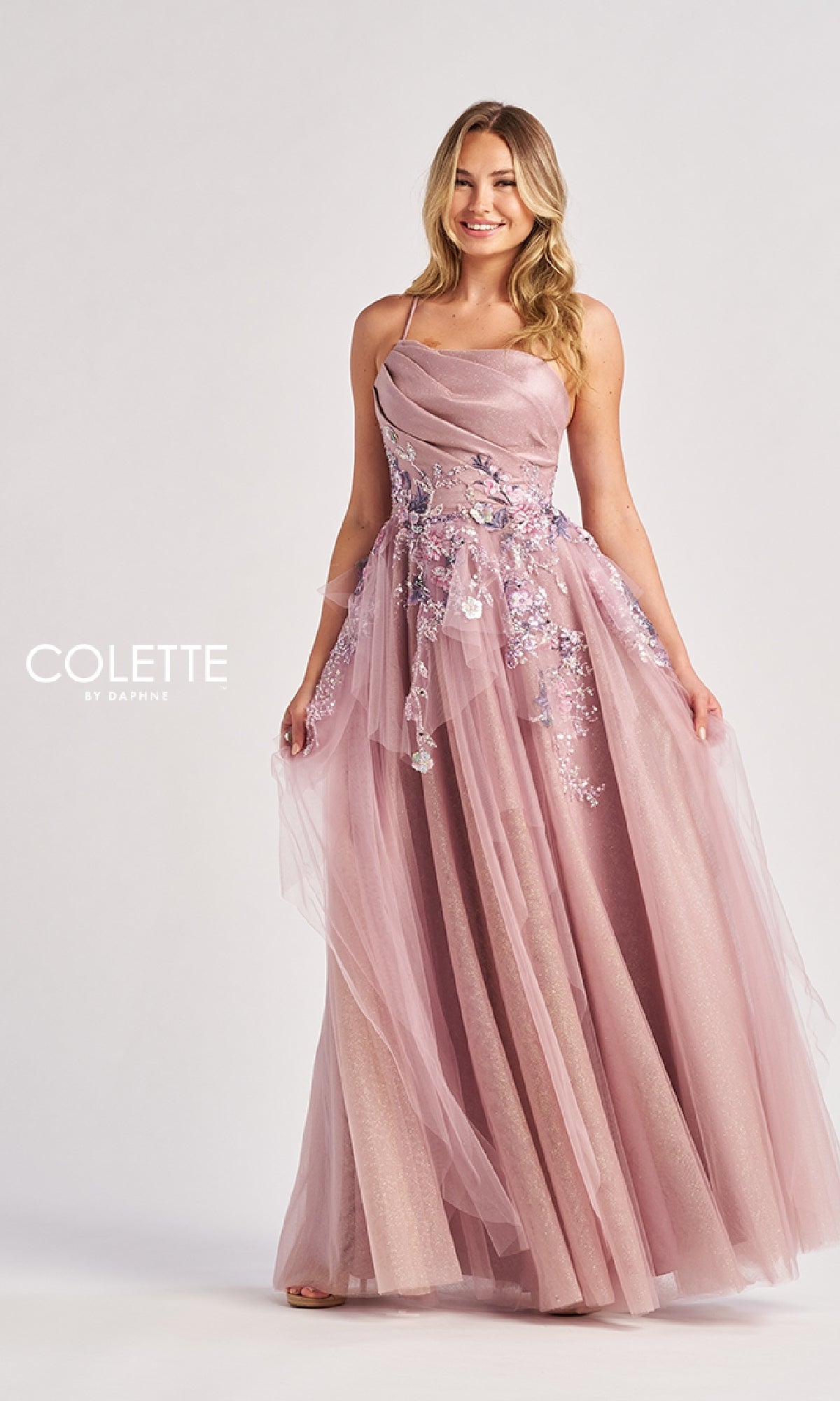 A woman wears the Colette CL8400 long prom dress, featuring a floor-length blush pink tulle A-line skirt, floral embroidery, thin straps, an embroidered waist, and a fitted ruched bodice.